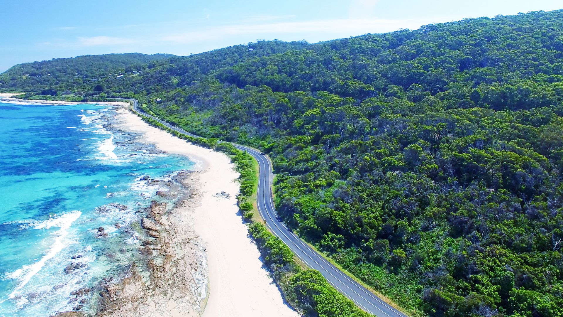 Aerial view of the Great Ocean Road winding between a beach and forested hills
